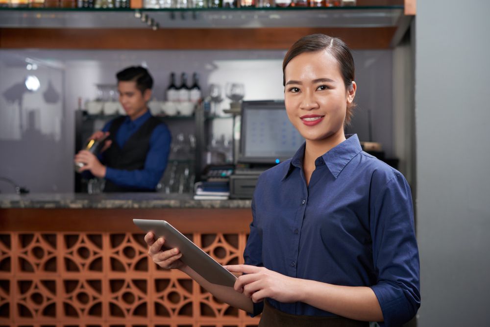 Manajer restoran tersenyum puas sambil memantau bisnisnya, menggambarkan kemudahan pengelolaan usaha dengan laporan real-time dari aplikasi kasir.